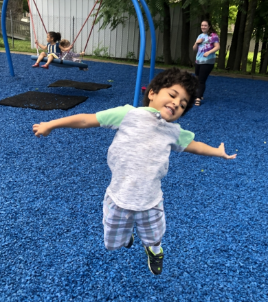 young boy at a playground jumping in the air with his arms out. there is blue playground mulch around him