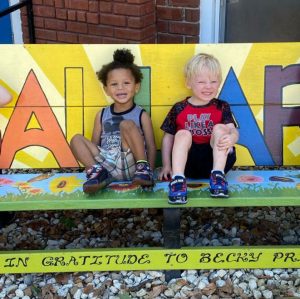 two young children sitting on a multi colored bench. both have their legs up and are smiling