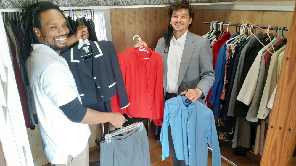 two men holding up suit jackets and shirts in front of racks of shoes and clothes