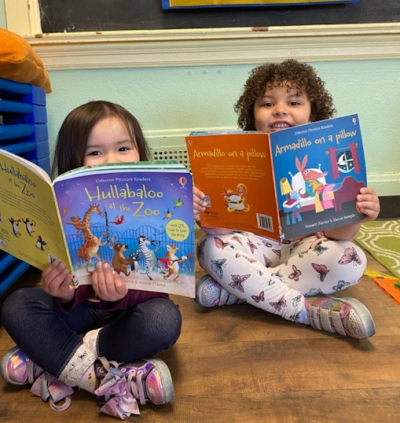 two young girls sitting on the floor of a classroom each reading a book